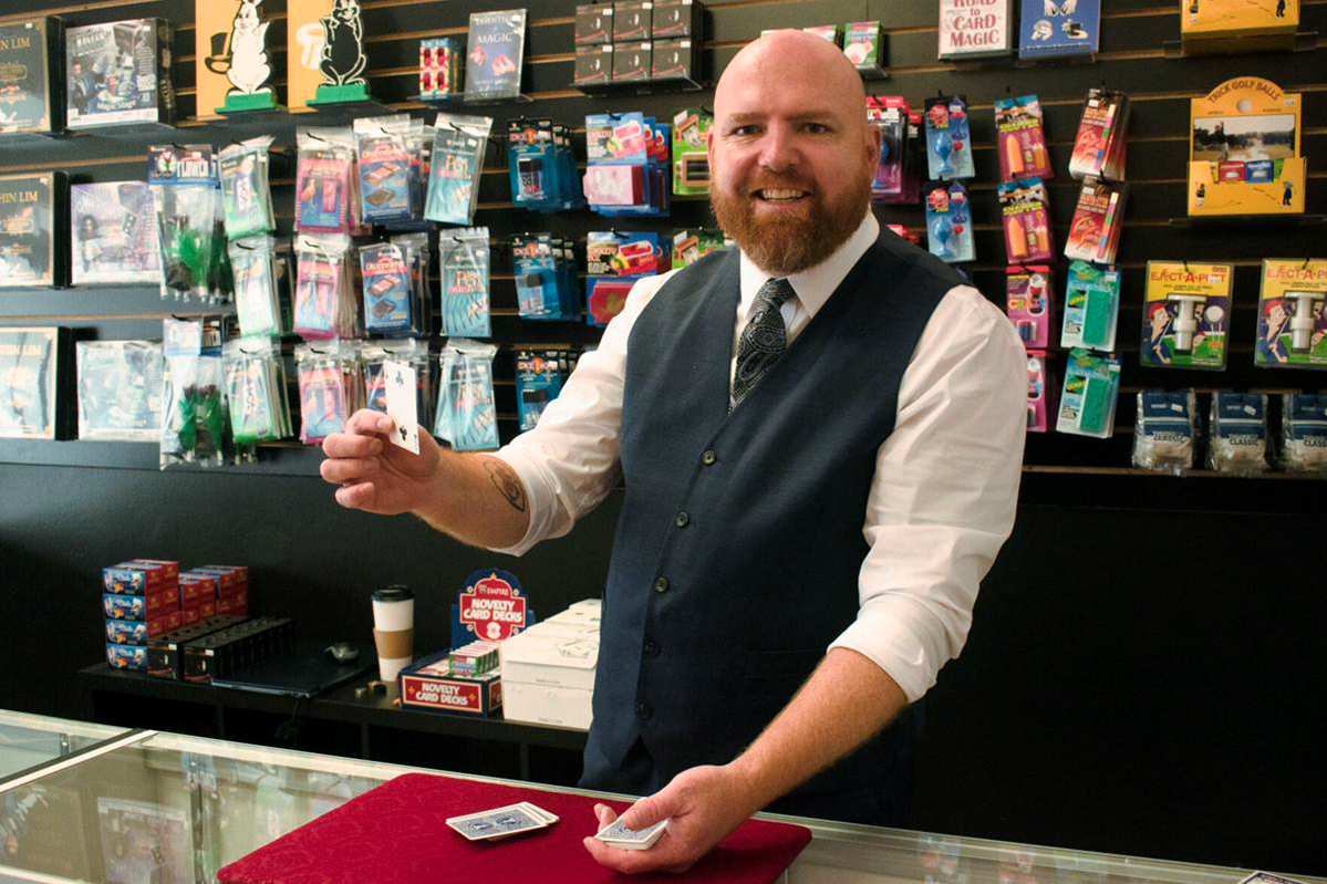 Seth Howard stands behind a counter holding a playing card in his right hand