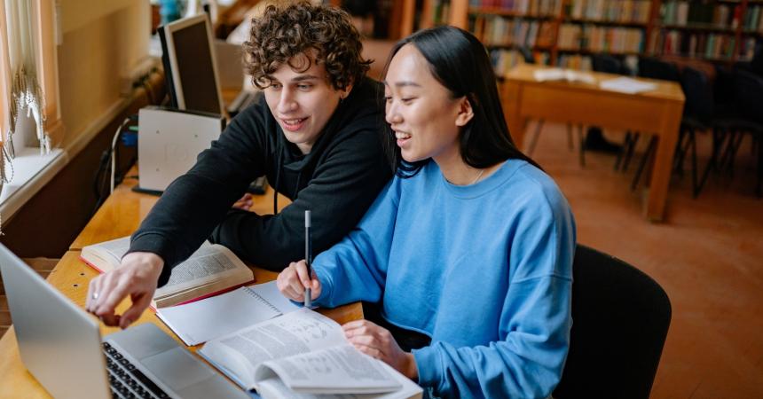 Two people look at a computer screen. One points excitedly to the screen while one of them takes notes.