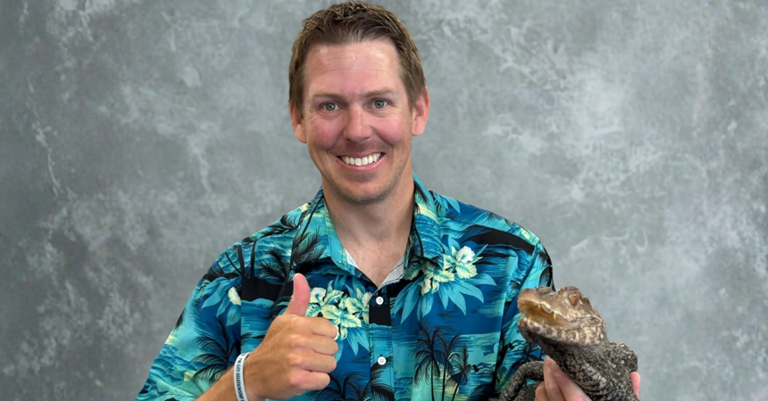 James McNamara, a light-skinned man with short brown hair and wearing a Hawaiian shirt, gives a thumbs up. In his other hand, he holds a small crocodile-like reptile.