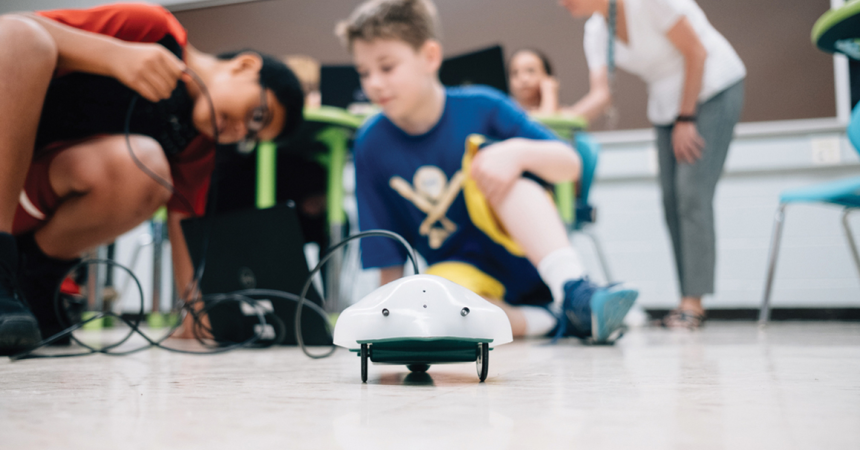Two children crouch over a Finch robot, a small disc-shaped device on wheels.