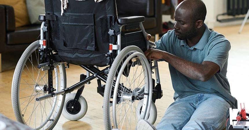 A dark-skinned man sits next to a wheelchair, examining a mechanism within.