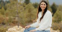 Whitney Wedam, a smiling woman with light skin and brown hair poses on a rock in front of a sagebrush landscape.