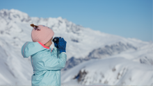 A young girl in a winter parka and pink hat holds up binoculars in a snowy landscape.