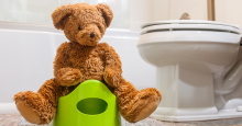 A teddy bear sits on a bright green training potty. An adult-sized toilet can be seen in the background.