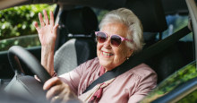 A white woman in sunglasses smiles and waves from the driver's seat of a car.