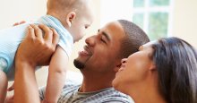 A Black man and woman cuddle and coo at a light-skinned infant.