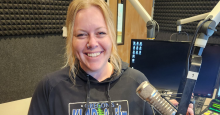 Melissa Tolbert, a light-skinned woman with long blond hair and wearing a KLAD station hoodie, smiles from inside a recording booth.
