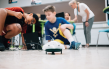 Two children crouch over a Finch robot, a small disc-shaped device on wheels.