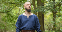 A bald, bearded man wears a medieval-era-inspired tunic in blue linen.